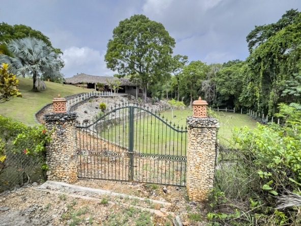 buyDRproperty Gated entrance to building lot with stone pillars, lush landscaping, and thatched roof building. Sosua Real Estate