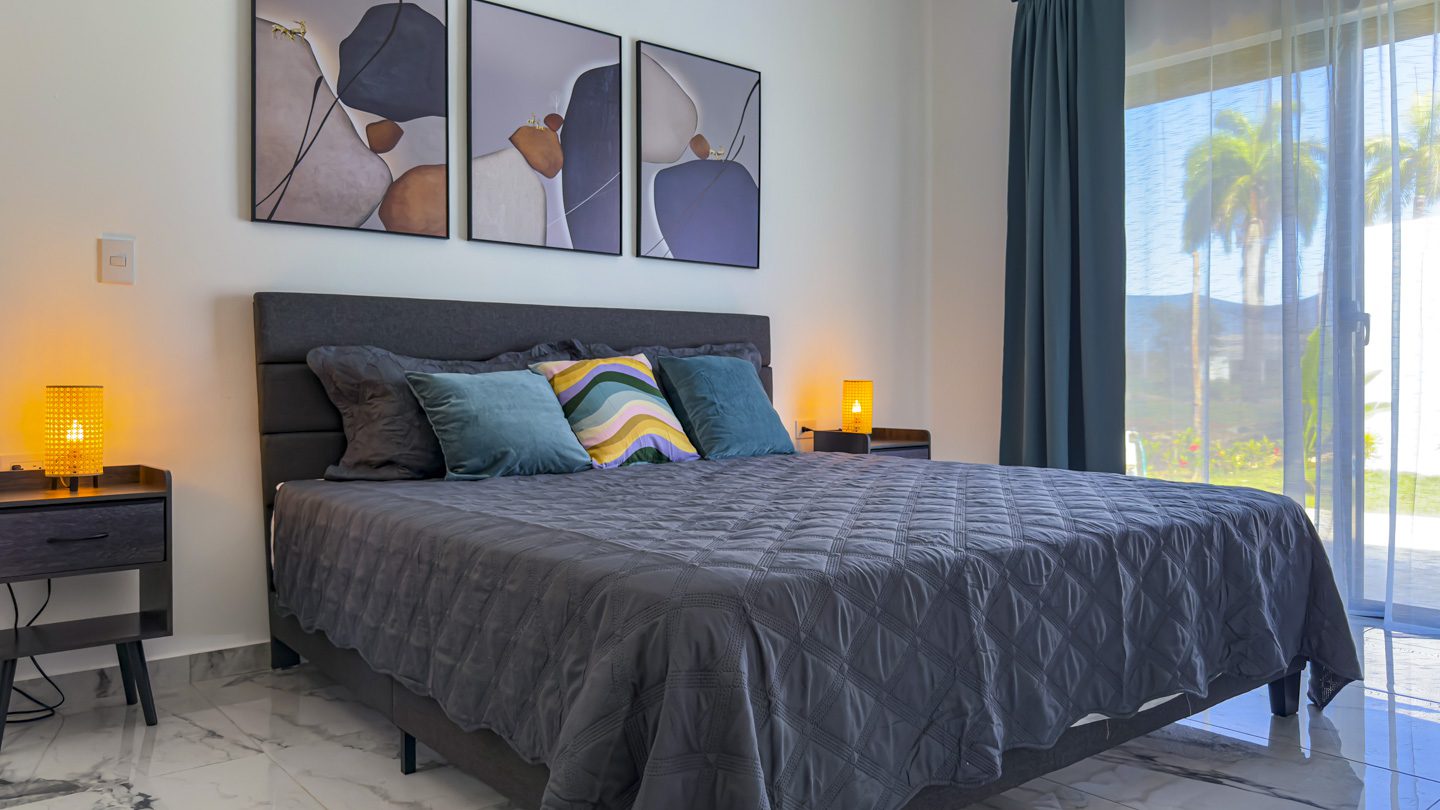 buyDRproperty Modern bedroom with a gray bed, bedside lamps, and artwork. View of palm trees. Sosua Real Estate