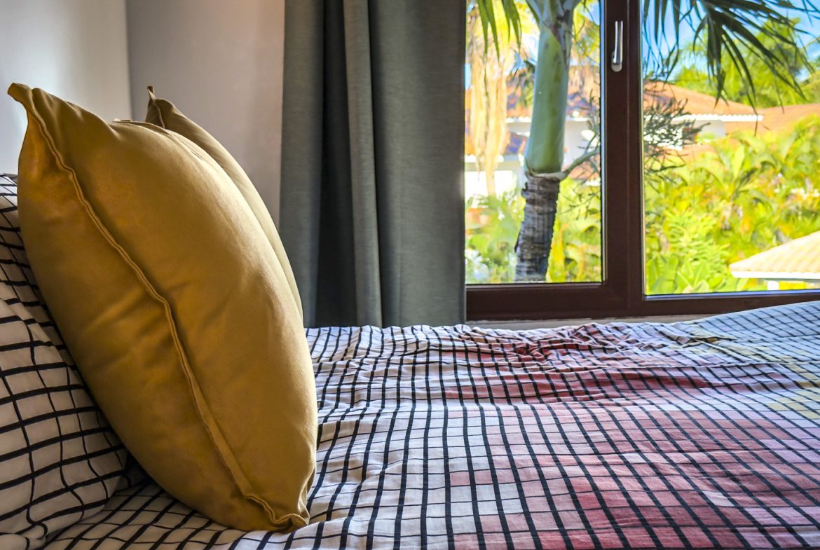 buyDRproperty Bedroom in a luxury villa with ocean views, featuring a bed with patterned linens and a window overlooking tropical foliage. Sosua Real Estate