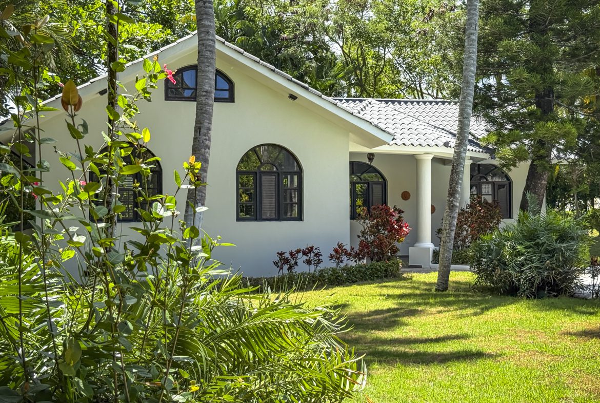 buyDRproperty White tropical house with arched windows, surrounded by lush greenery and palm trees. Sosua Real Estate