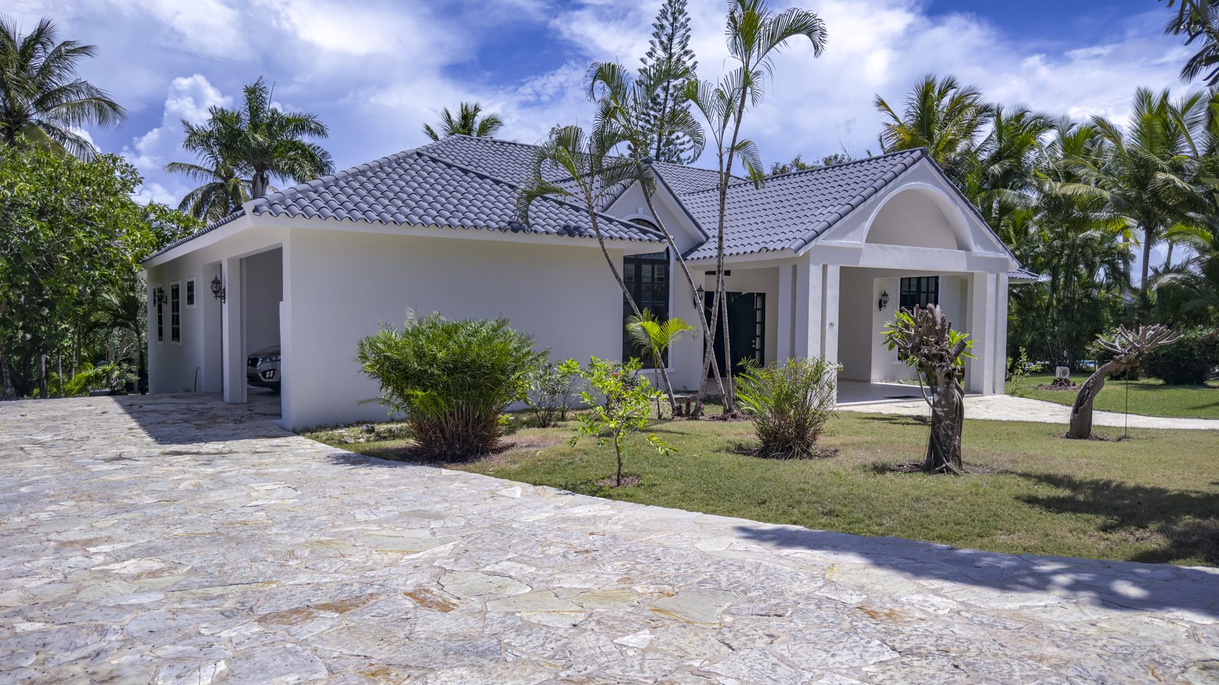 buyDRproperty White tropical home with gray tile roof and stone driveway, surrounded by palm trees Sosua Real Estate