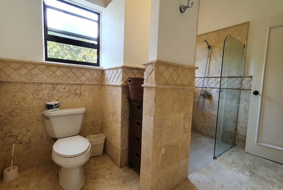 buyDRproperty Travertine tile bathroom with toilet, shower, and natural light from window. Sosua Real Estate