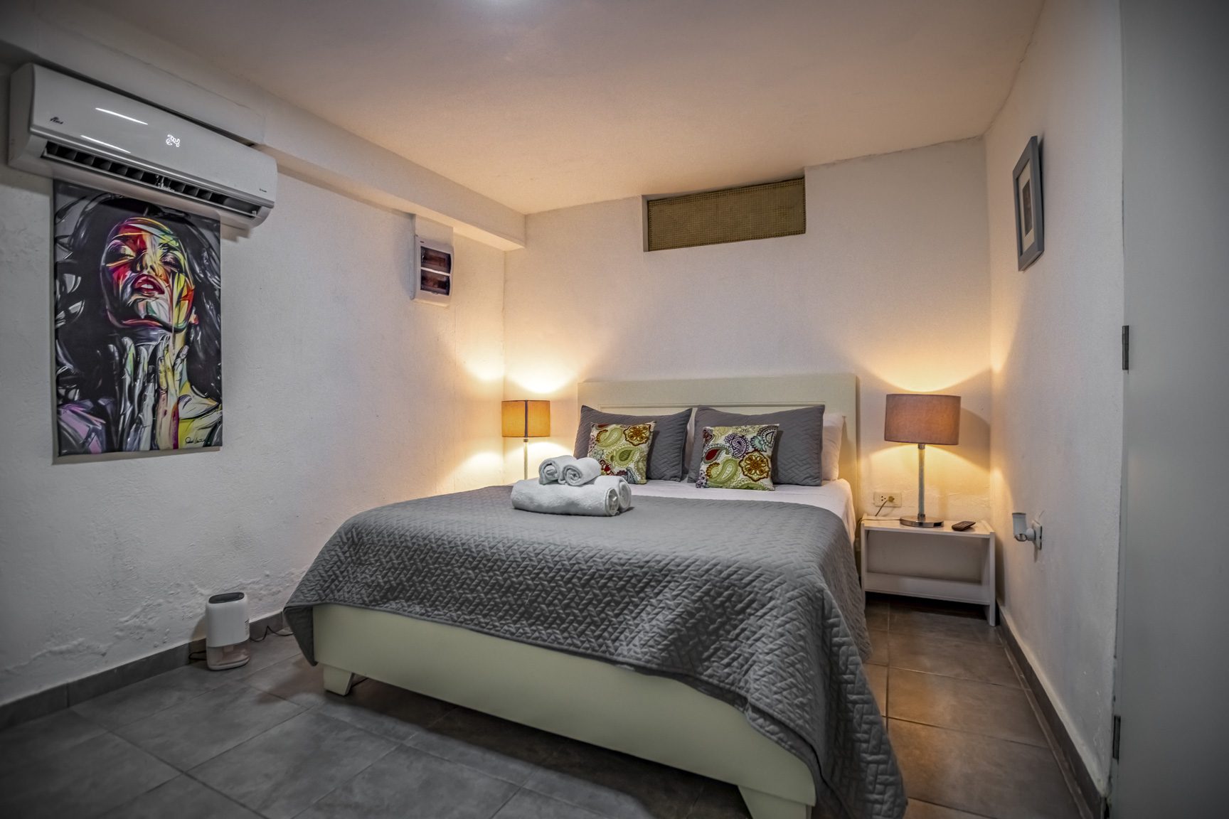 buyDRproperty Cozy bedroom with queen bed, gray quilt, bedside lamps, and modern wall art. Sosua Real Estate