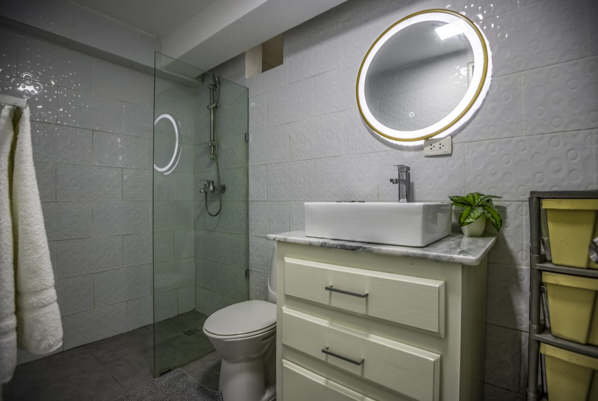 buyDRproperty Modern bathroom with white square tiles, round mirror, vessel sink, and glass shower enclosure. Sosua Real Estate
