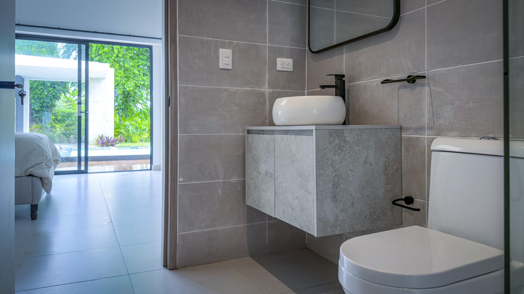 buyDRproperty Modern bathroom with gray tile, white sink, toilet, and view of the luxury villa's pool area. Sosua Real Estate