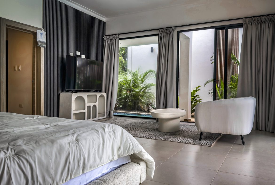 buyDRproperty Luxury villa bedroom with TV, seating area, and patio view of lush greenery and pool. Sosua Real Estate