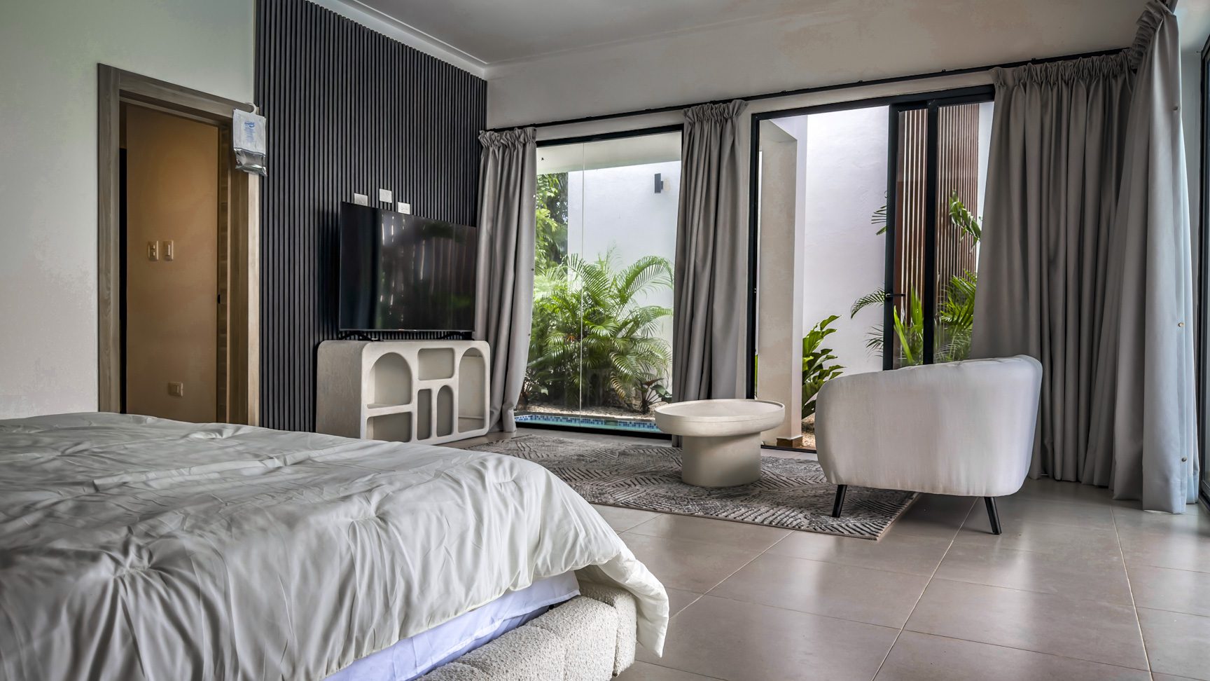 buyDRproperty Luxury villa bedroom with TV, seating area, and patio view of lush greenery and pool. Sosua Real Estate