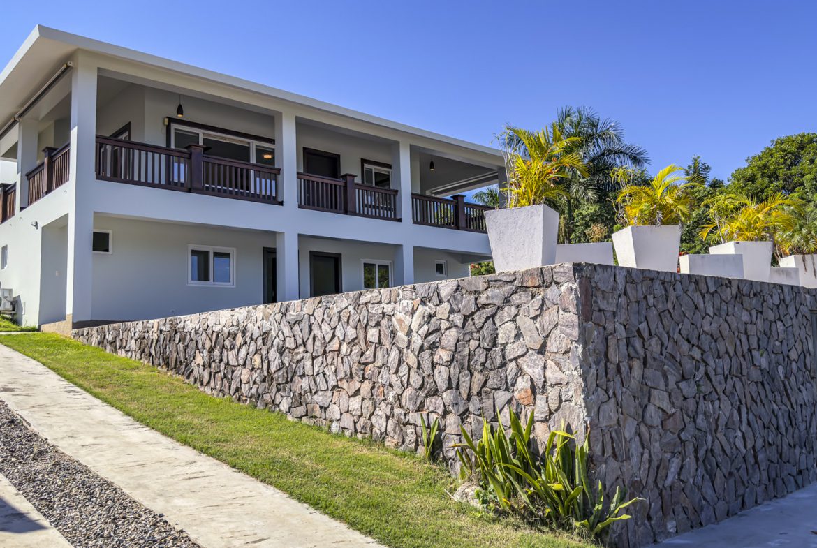 buyDRproperty La Mulata villa for sale: Exterior view of a modern 3-bedroom villa with a stone retaining wall and a covered balcony. Sosua Real Estate
