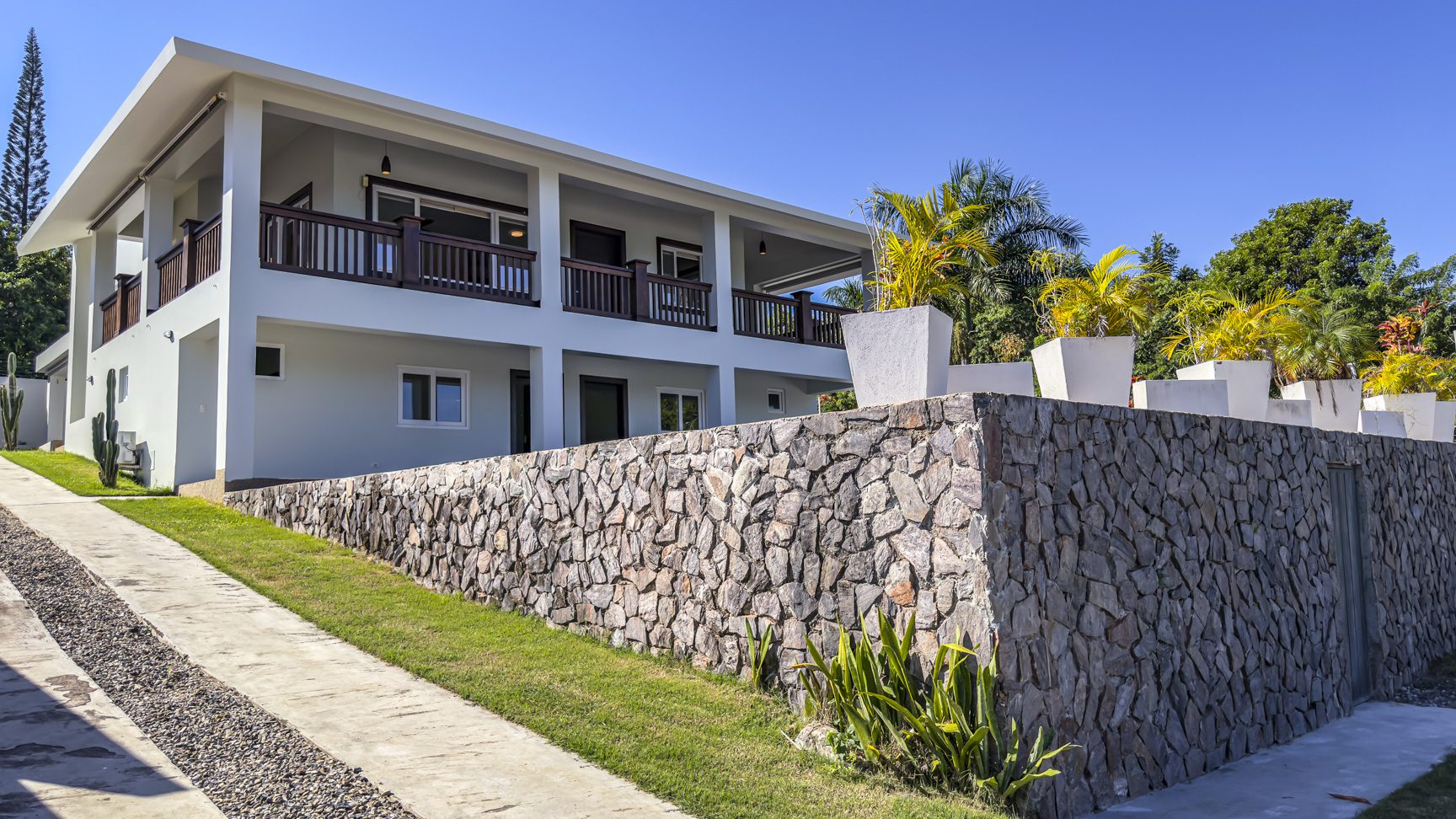 buyDRproperty La Mulata villa for sale: Exterior view of a modern 3-bedroom villa with a stone retaining wall and a covered balcony. Sosua Real Estate