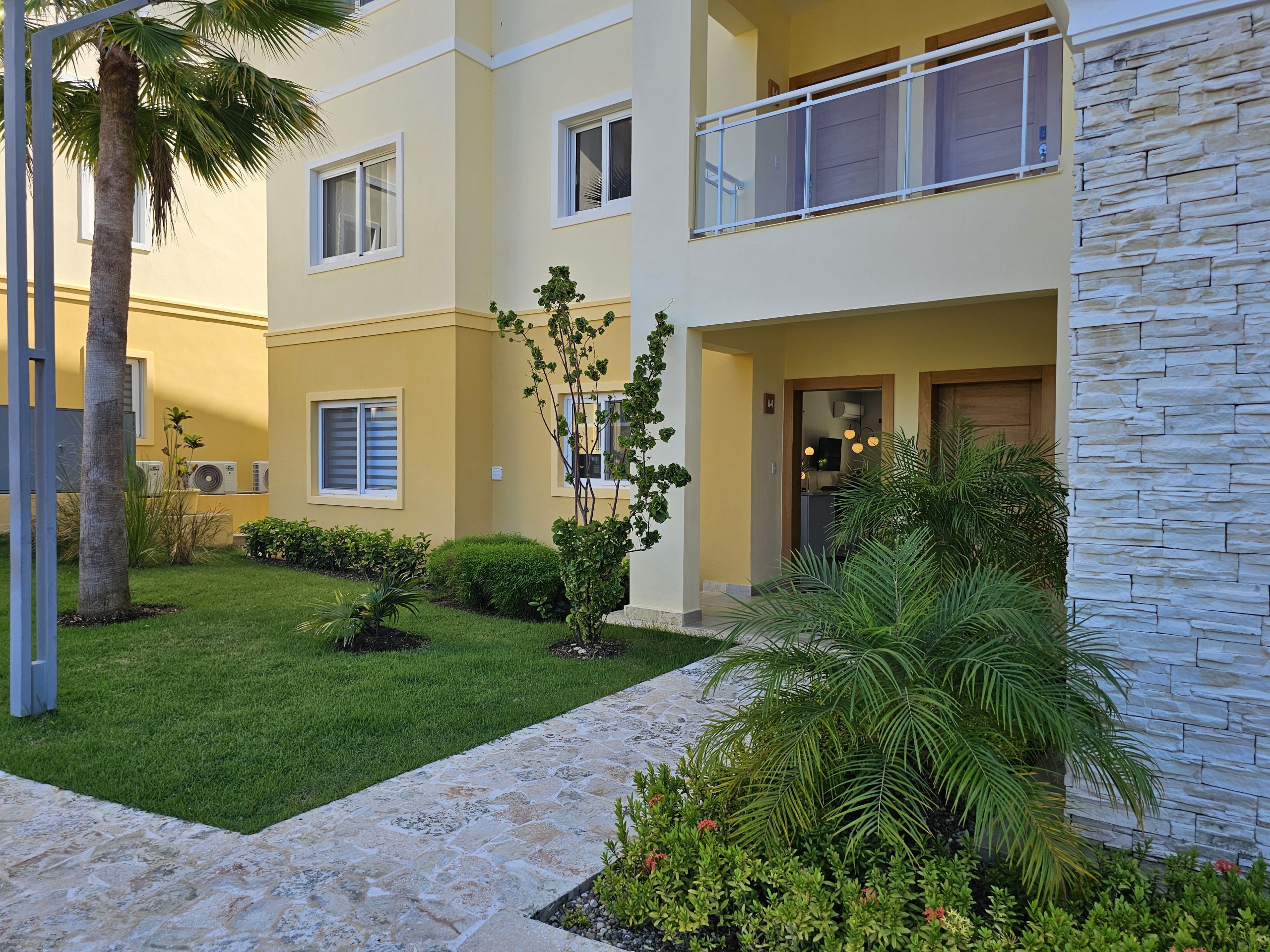 buyDRproperty Exterior of a modern yellow apartment building with lush green lawn and stone walkway. Sosua Real Estate