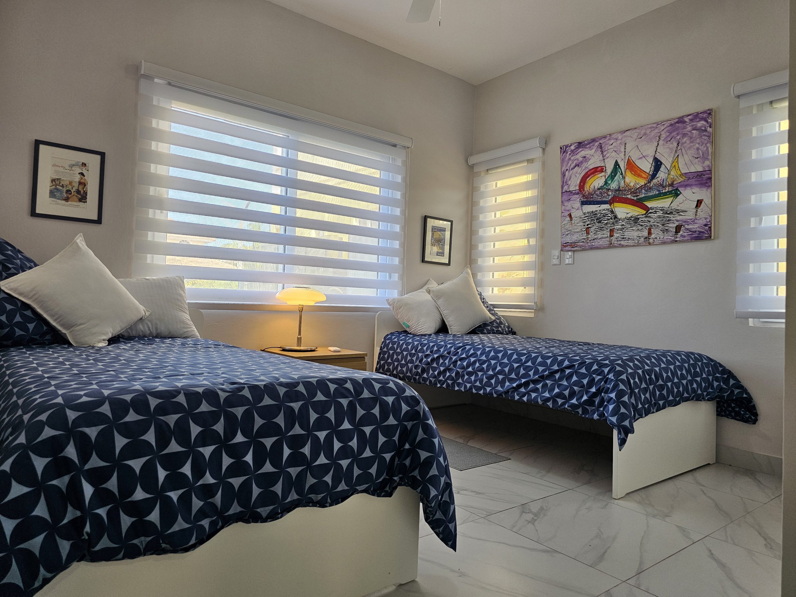 buyDRproperty Bright bedroom with two twin beds, blue patterned bedding, colorful sailboat painting, and white tile floor. Sosua Real Estate