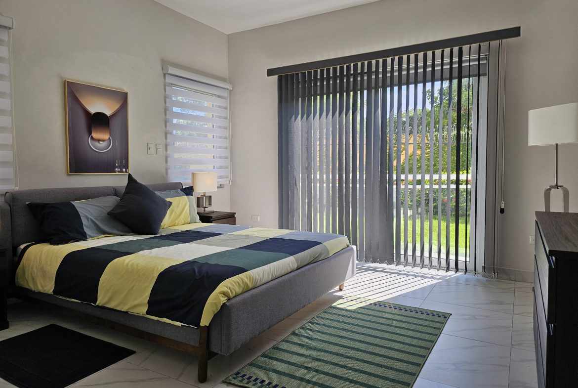 buyDRproperty Bedroom with modern gray bed, colorful quilt, and bright sunlight streaming through vertical blinds. Sosua Real Estate