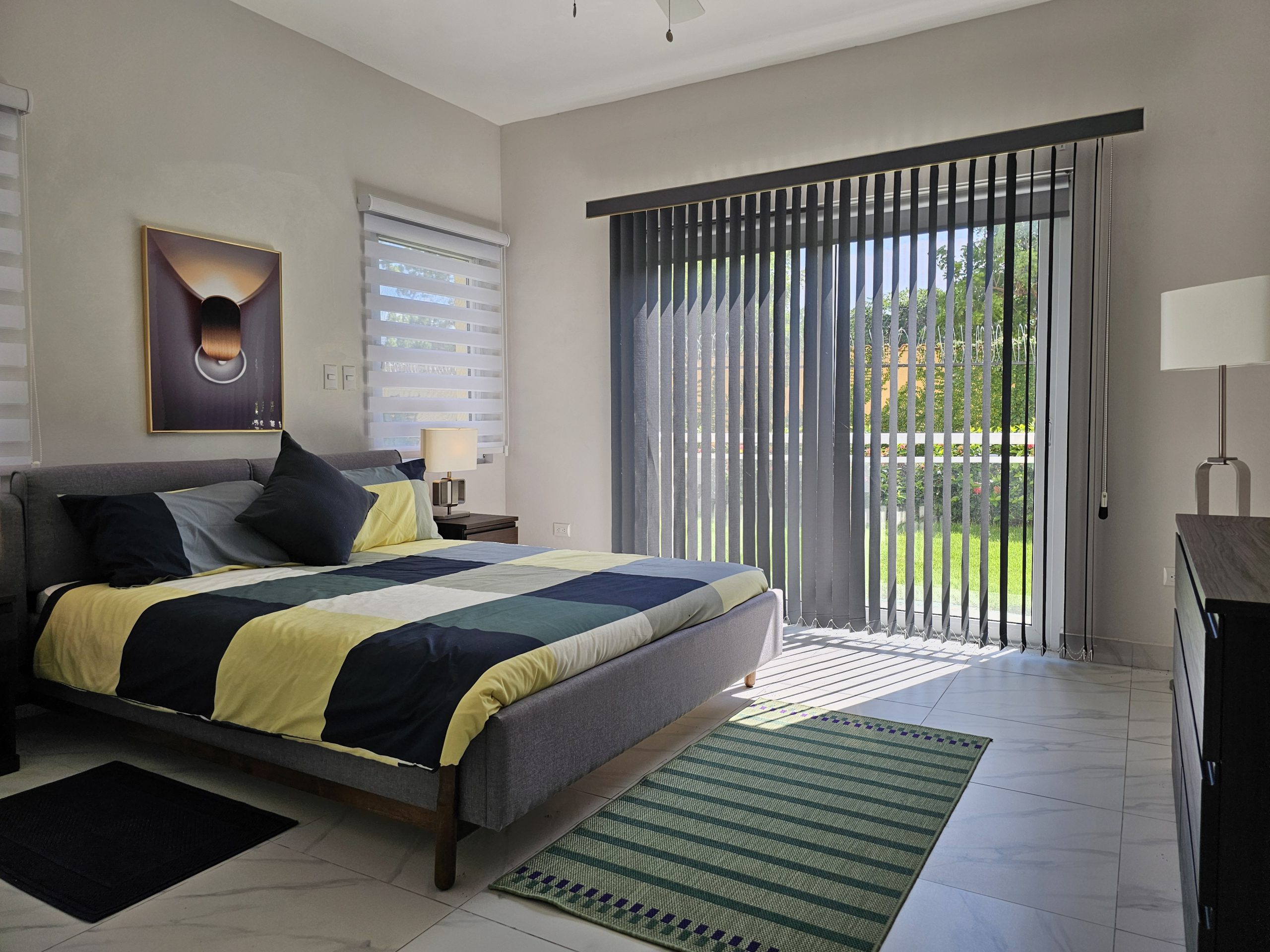 buyDRproperty Bedroom with modern gray bed, colorful quilt, and bright sunlight streaming through vertical blinds. Sosua Real Estate