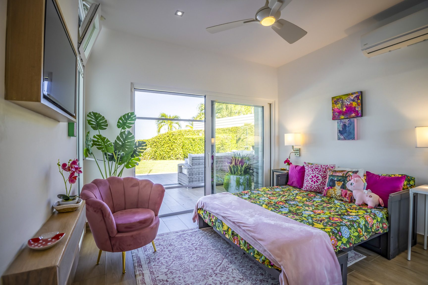 buyDRproperty Bright bedroom with floral bedding, pink accents, open patio doors to garden view, and a pink velvet chair. Sosua Real Estate