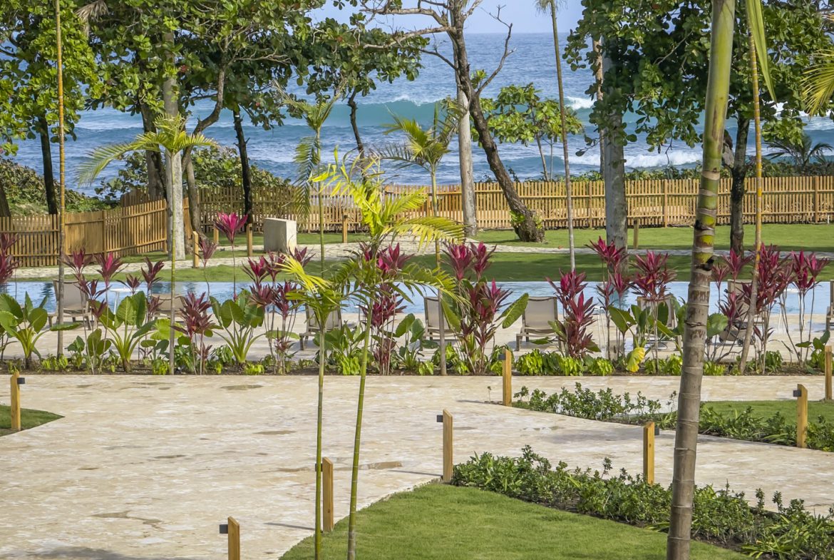 buyDRproperty Tropical resort view with pool, palm trees, and ocean backdrop. Lush landscaping and sandy pathway. Sosua Real Estate