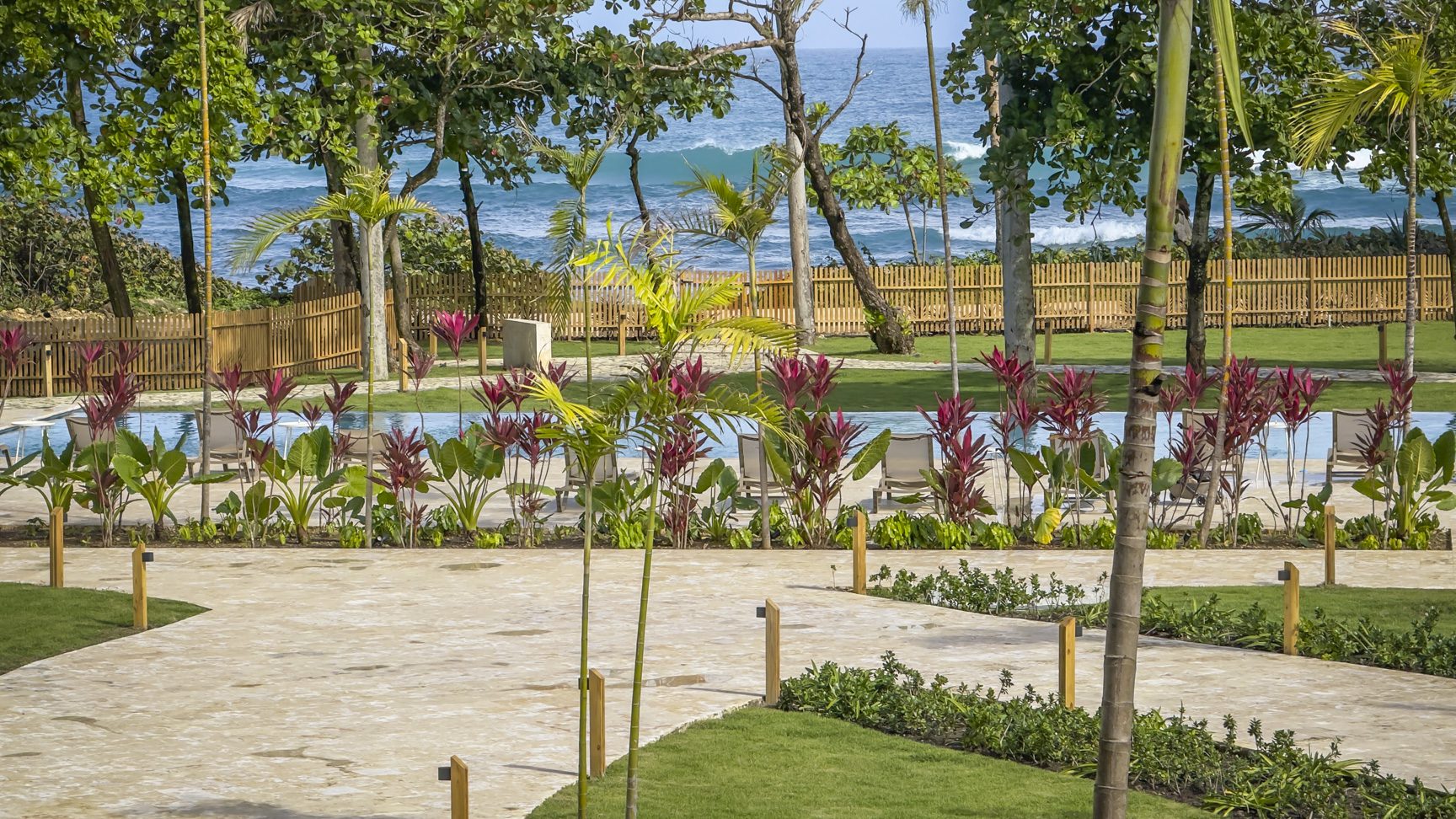 buyDRproperty Tropical resort view with pool, palm trees, and ocean backdrop. Lush landscaping and sandy pathway. Sosua Real Estate