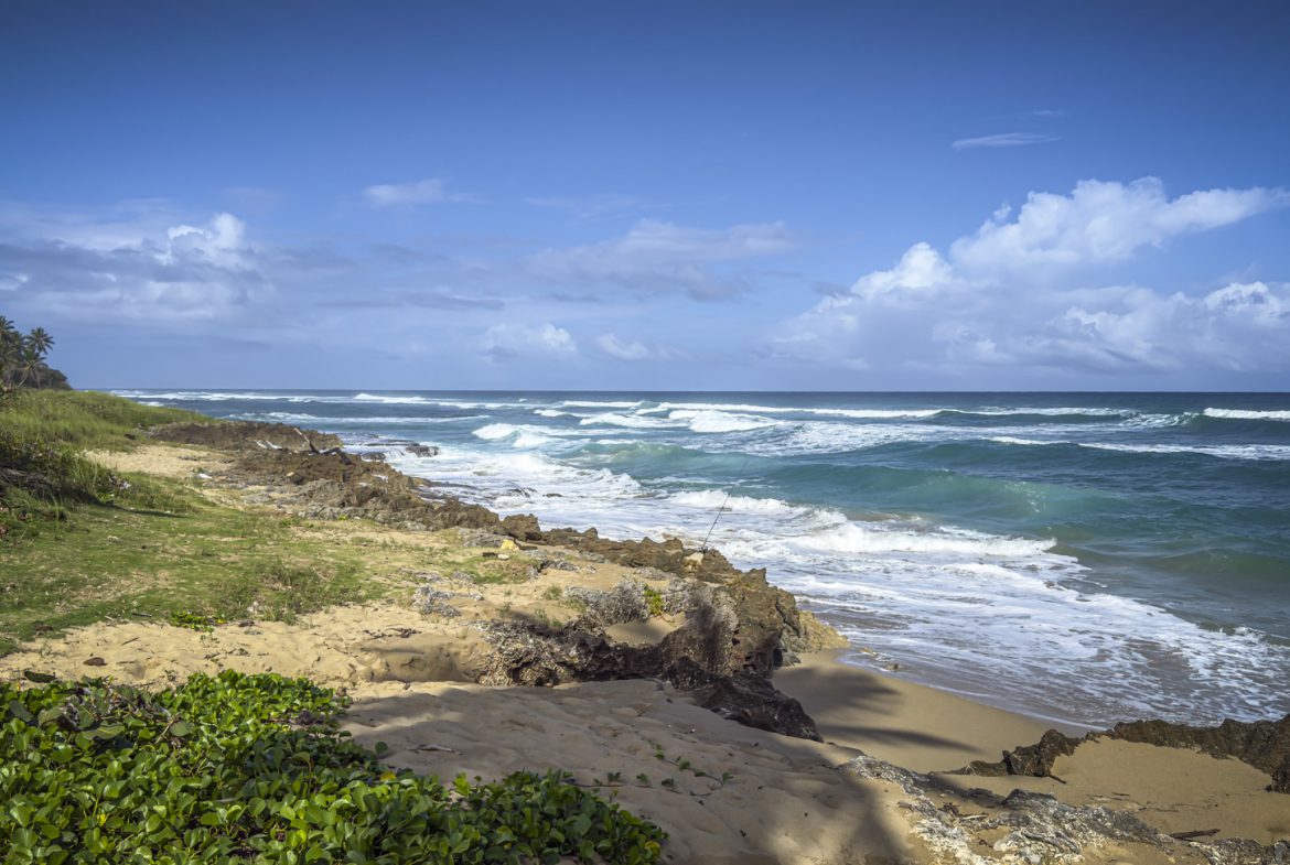 buyDRproperty Turquoise waves crashing on a sandy beach with rocky outcrops and lush green vegetation under a blue sky. Sosua Real Estate