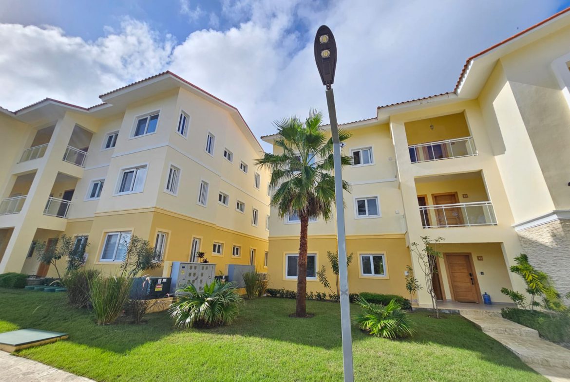 buyDRproperty Modern yellow apartment building with palm tree and green lawn on a sunny day. Sosua Real Estate