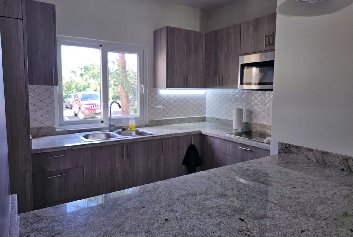 buyDRproperty Modern kitchen with granite countertops, wood cabinets, stainless steel microwave, and a bright window view. Sosua Real Estate