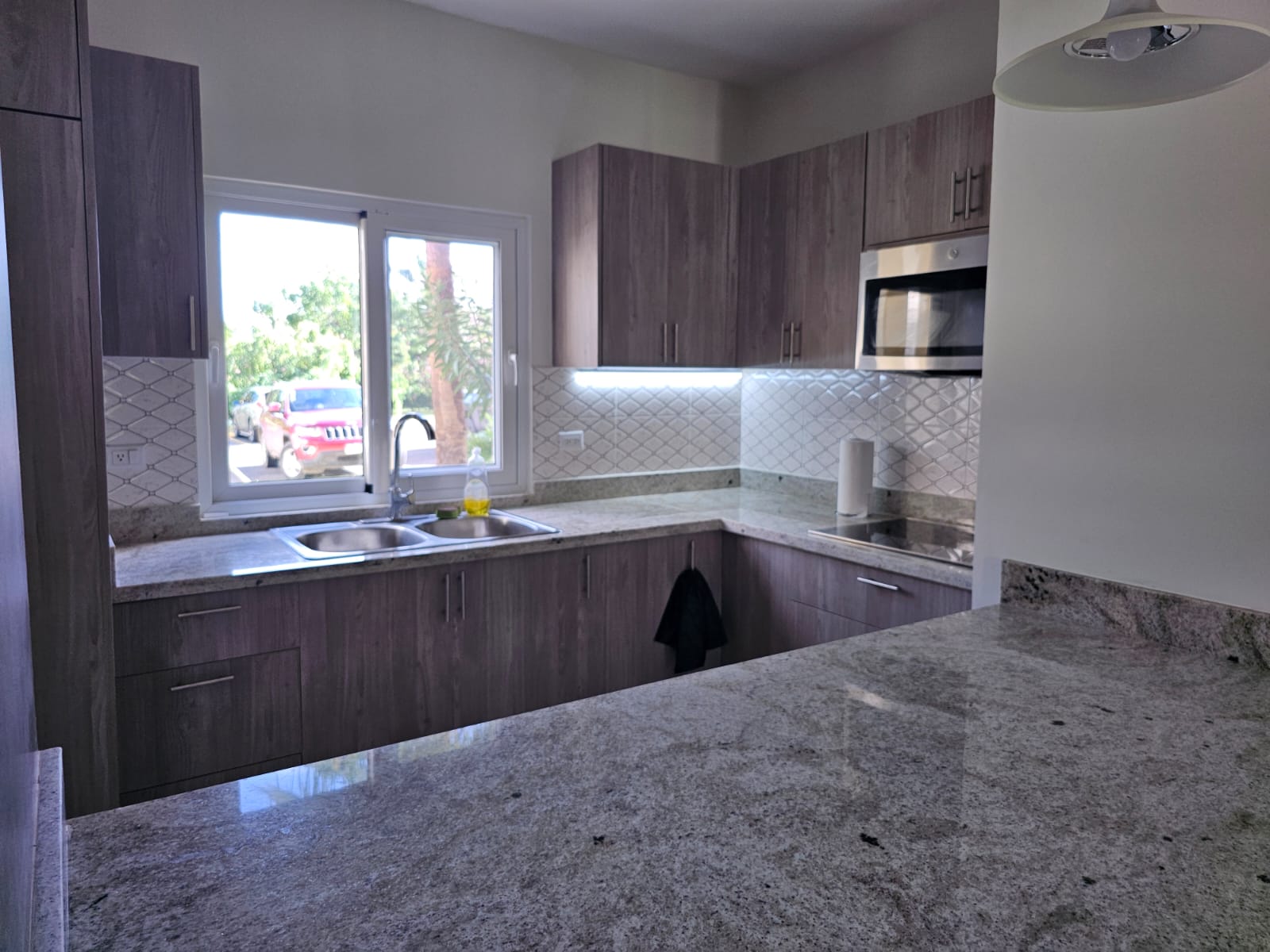 buyDRproperty Modern kitchen with granite countertops, wood cabinets, stainless steel microwave, and a bright window view. Sosua Real Estate