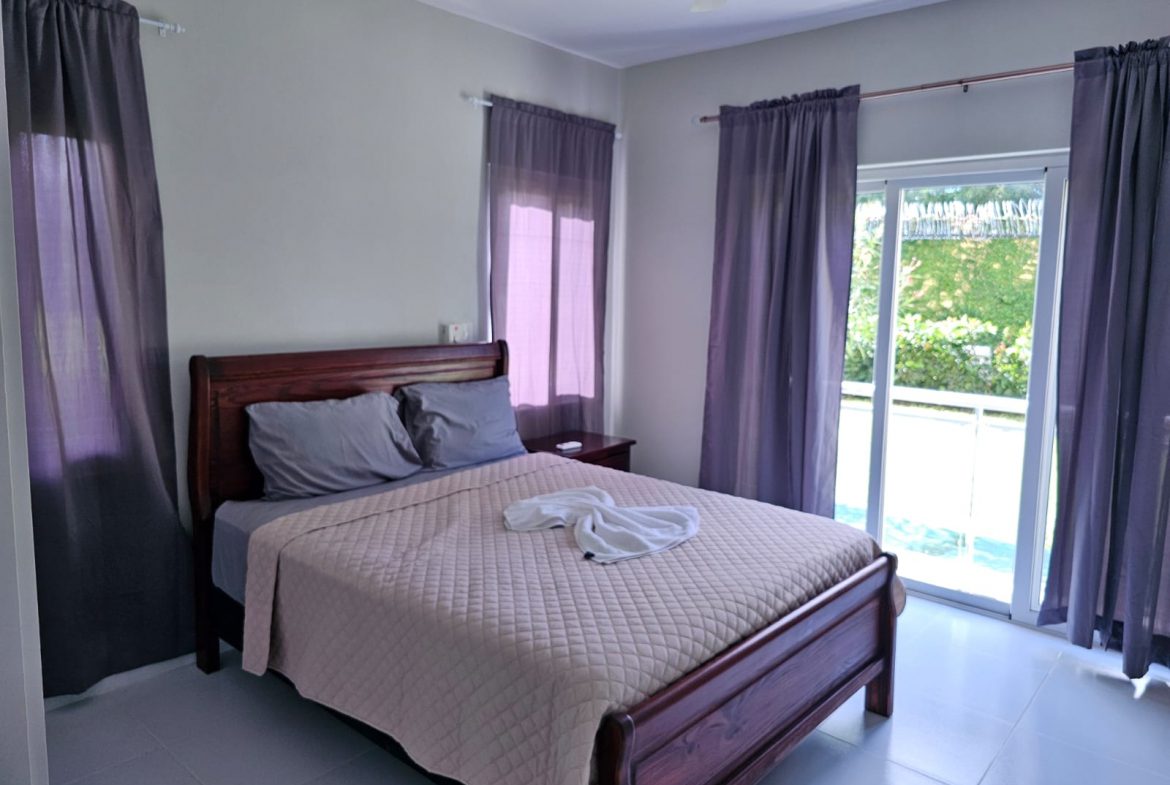 buyDRproperty Bedroom with a wooden bed, gray bedding, and purple curtains, leading to a balcony. Sosua Real Estate