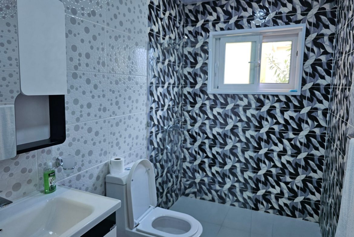 buyDRproperty Modern bathroom with white toilet, sink, and black/white patterned shower tiles, illuminated by natural light. Sosua Real Estate