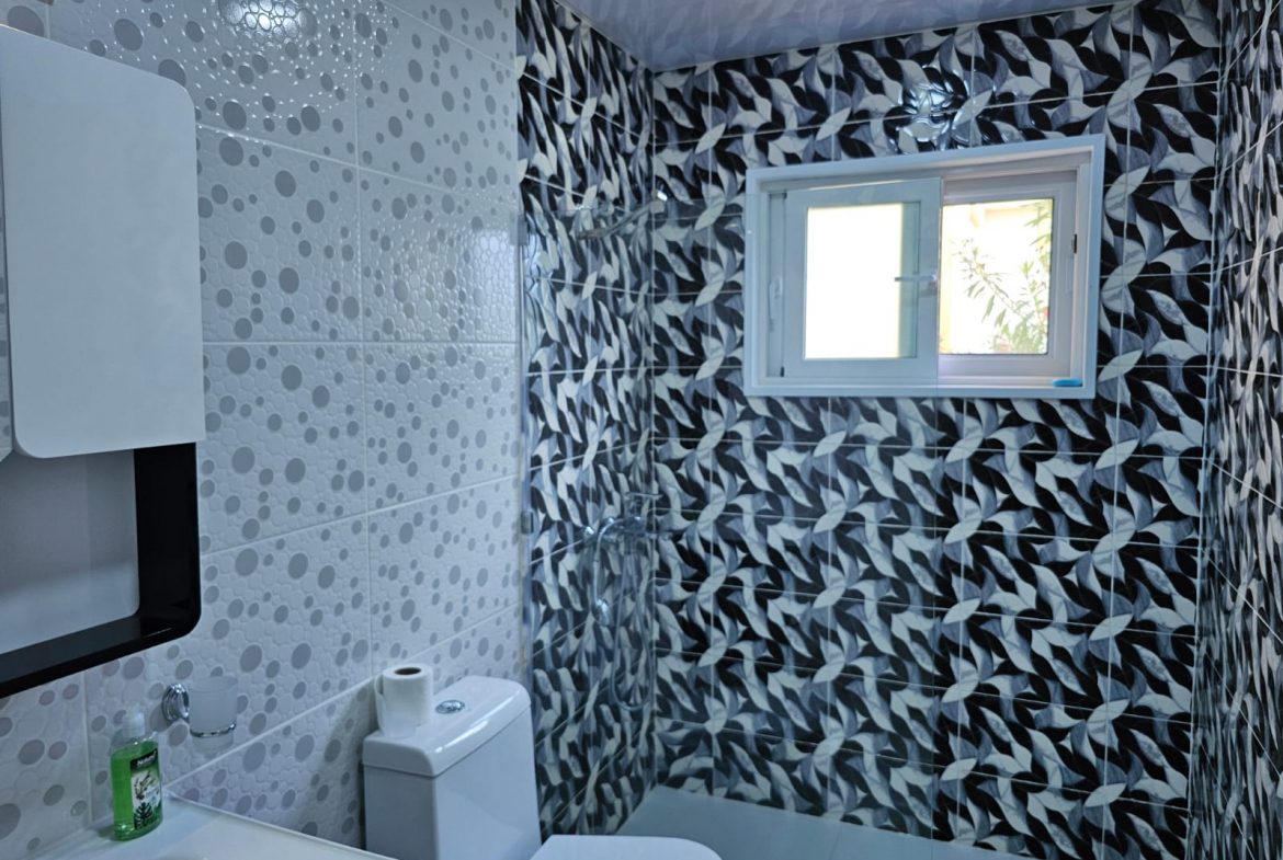 buyDRproperty Modern bathroom with gray patterned tiles, white toilet, sink, and window providing natural light. Sosua Real Estate