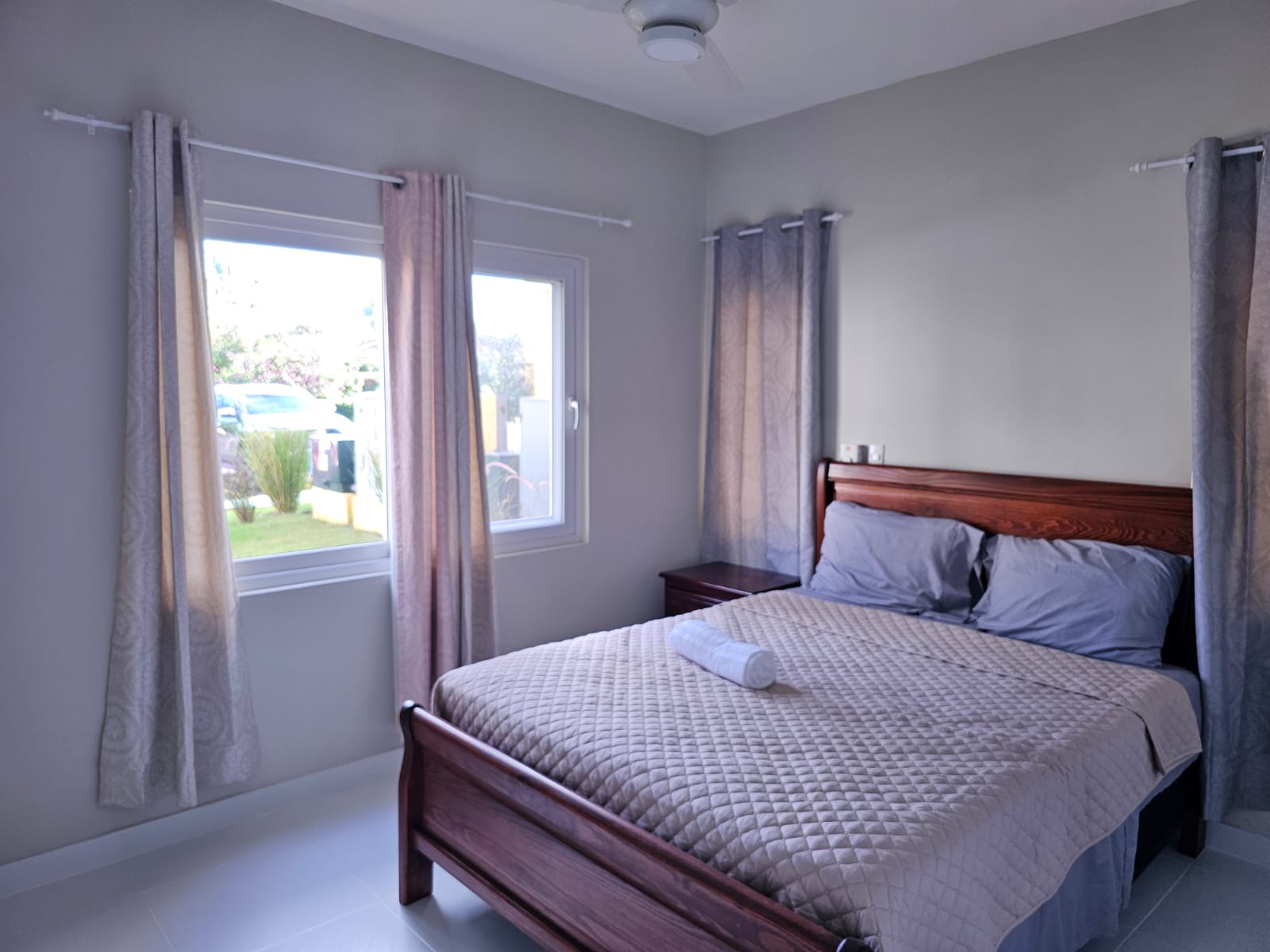 buyDRproperty Cozy bedroom with wooden bed frame, quilted beige bedspread, and two windows with curtains. Sosua Real Estate