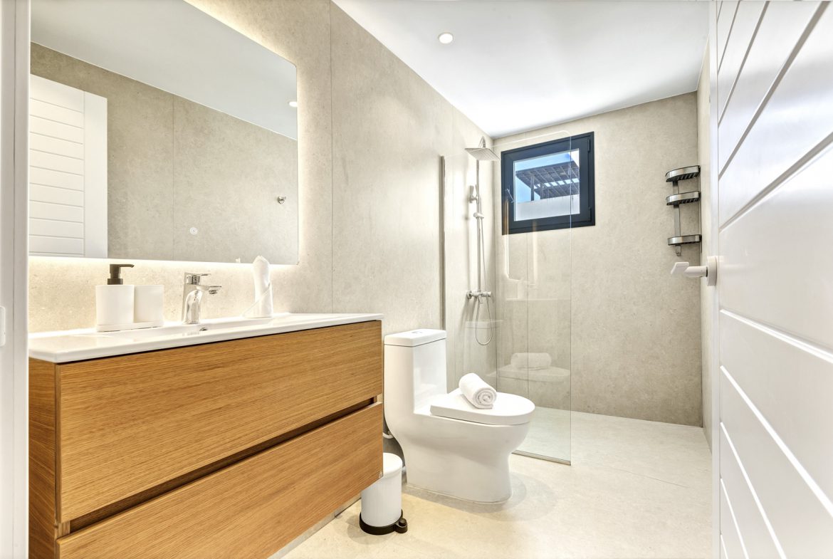 buyDRproperty Modern bathroom with wood vanity, toilet, glass shower, light tile, and white door. Sosua Real Estate