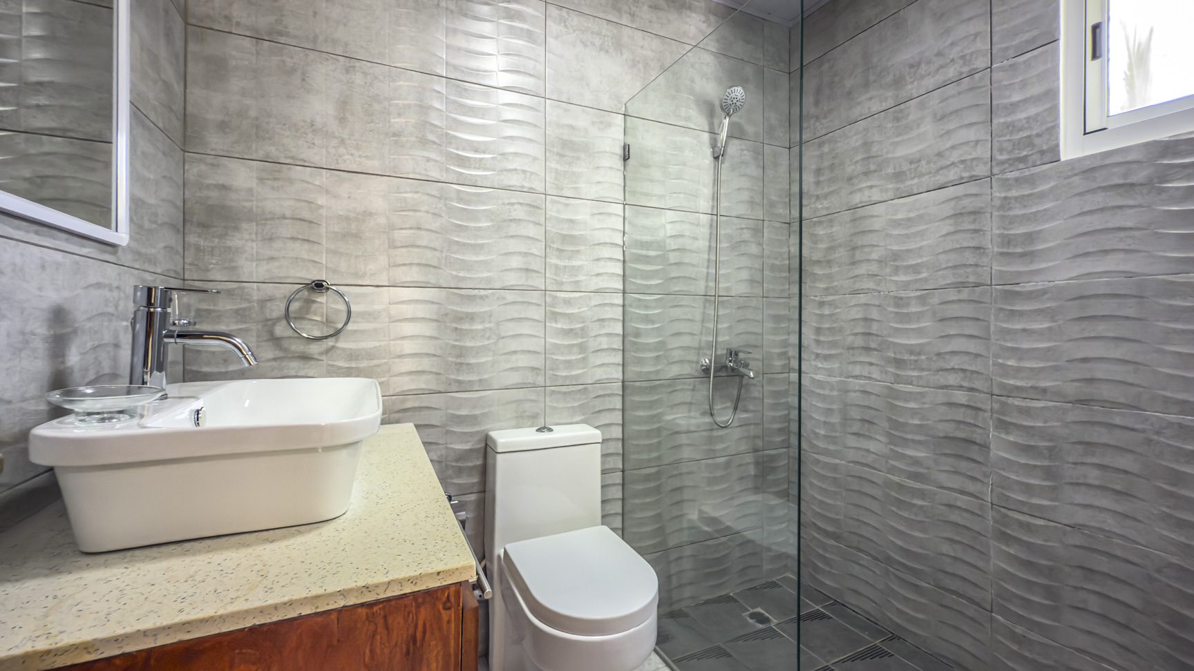 bathroom with textured gray tiles, white toilet, sink, and glass shower enclosure. Sosua Real Estate