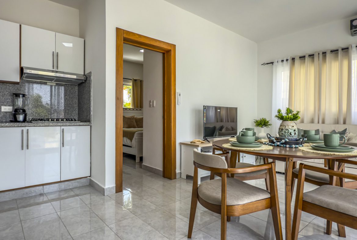 Modern apartment interior with kitchen, dining area, living room, and bedroom visible through doorway.