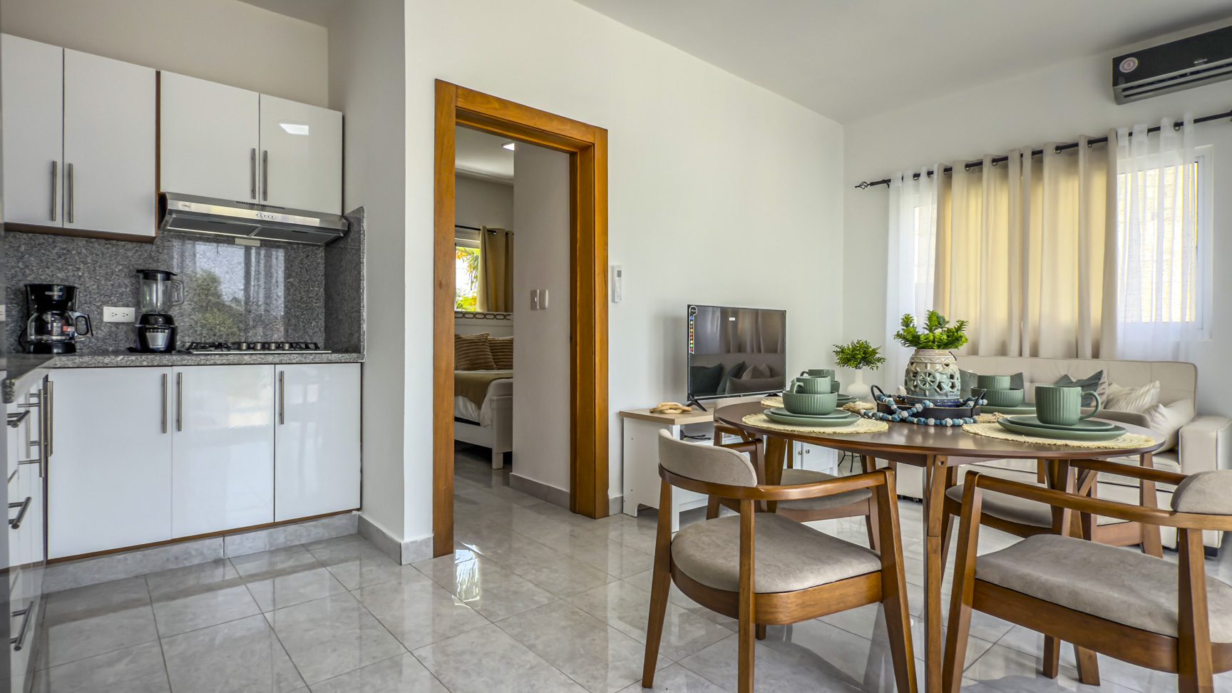 Modern apartment interior with kitchen, dining area, living room, and bedroom visible through doorway.