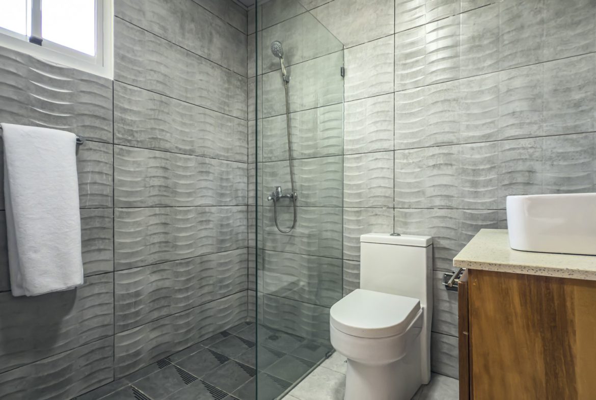 bathroom with textured gray tile, glass shower, toilet, and white sink on wood cabinet.