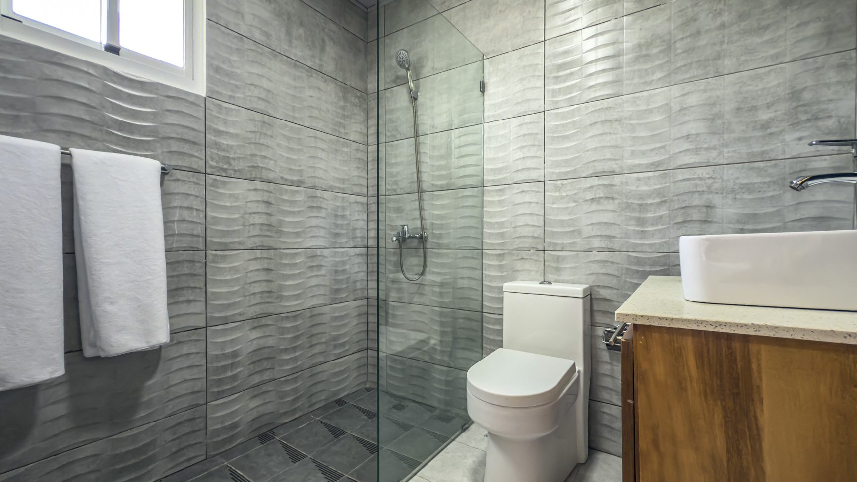 bathroom with textured gray tile, glass shower, toilet, and white sink on wood cabinet.