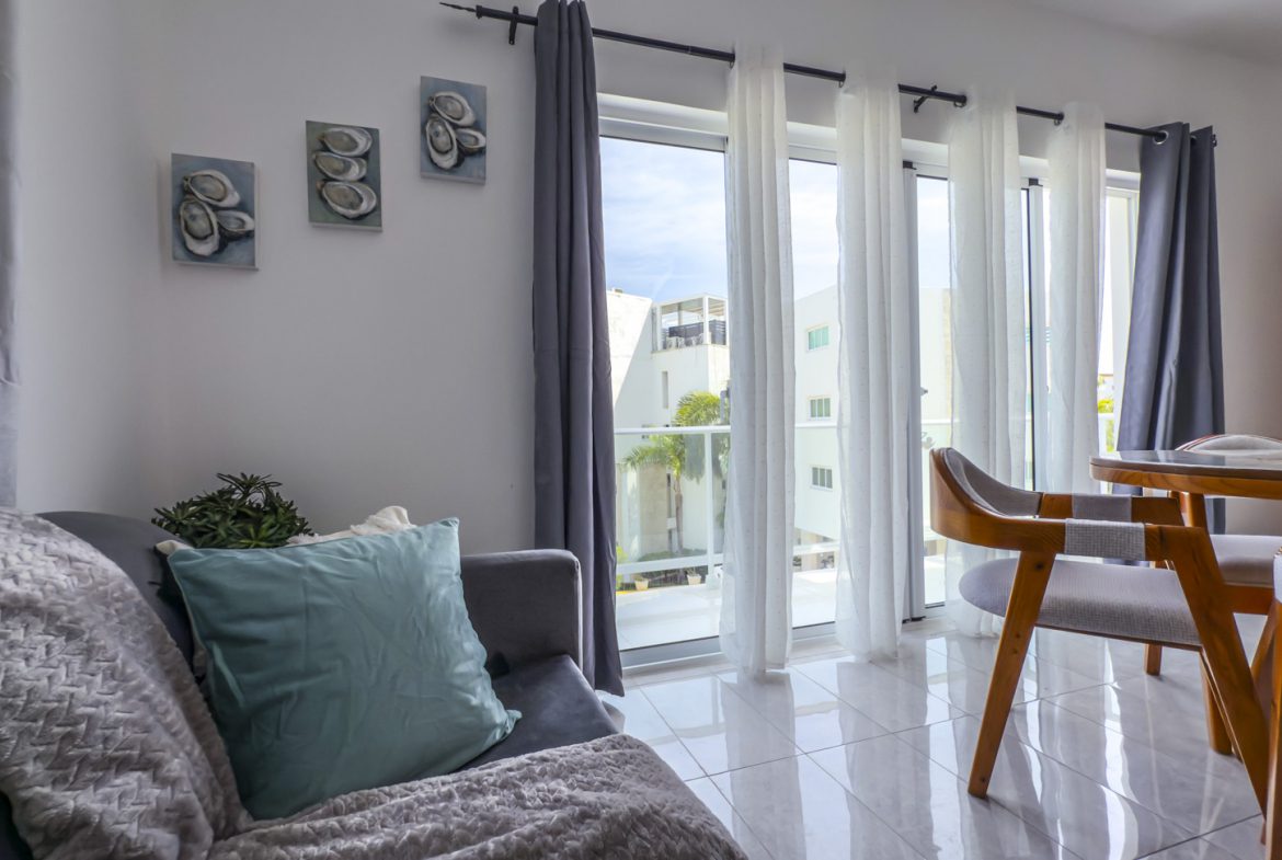 Bright living room with gray sofa, teal pillow, and dining area by window with white and gray curtains.