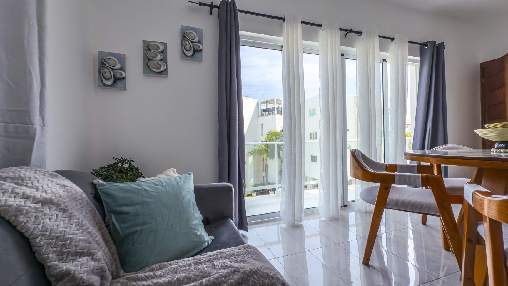 Bright living room with gray sofa, teal pillow, and dining area by window with white and gray curtains.