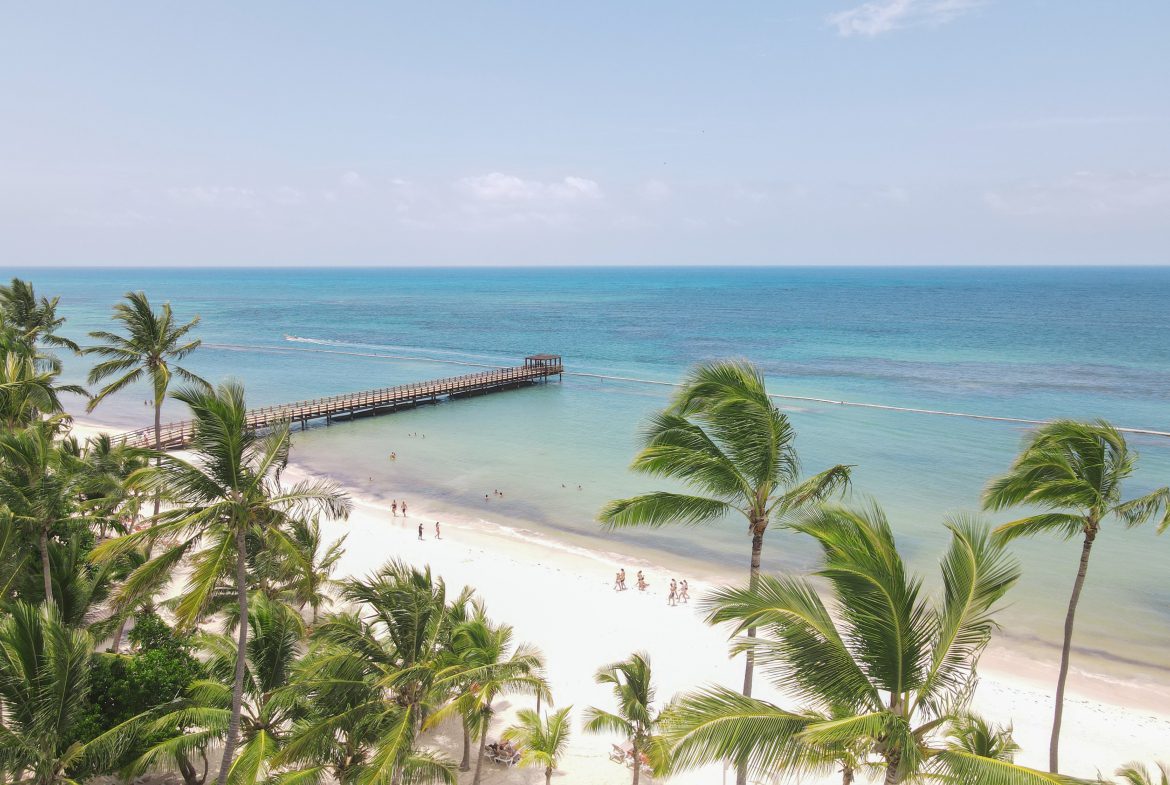 buyDRproperty Palm trees line a white sand beach with a pier extending into turquoise water. Sosua Real Estate