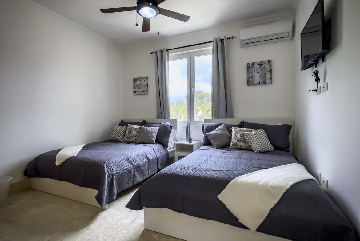 buyDRproperty Bedroom with two beds, gray bedding, ceiling fan, and wall-mounted TV. Neutral decor. Sosua Real Estate