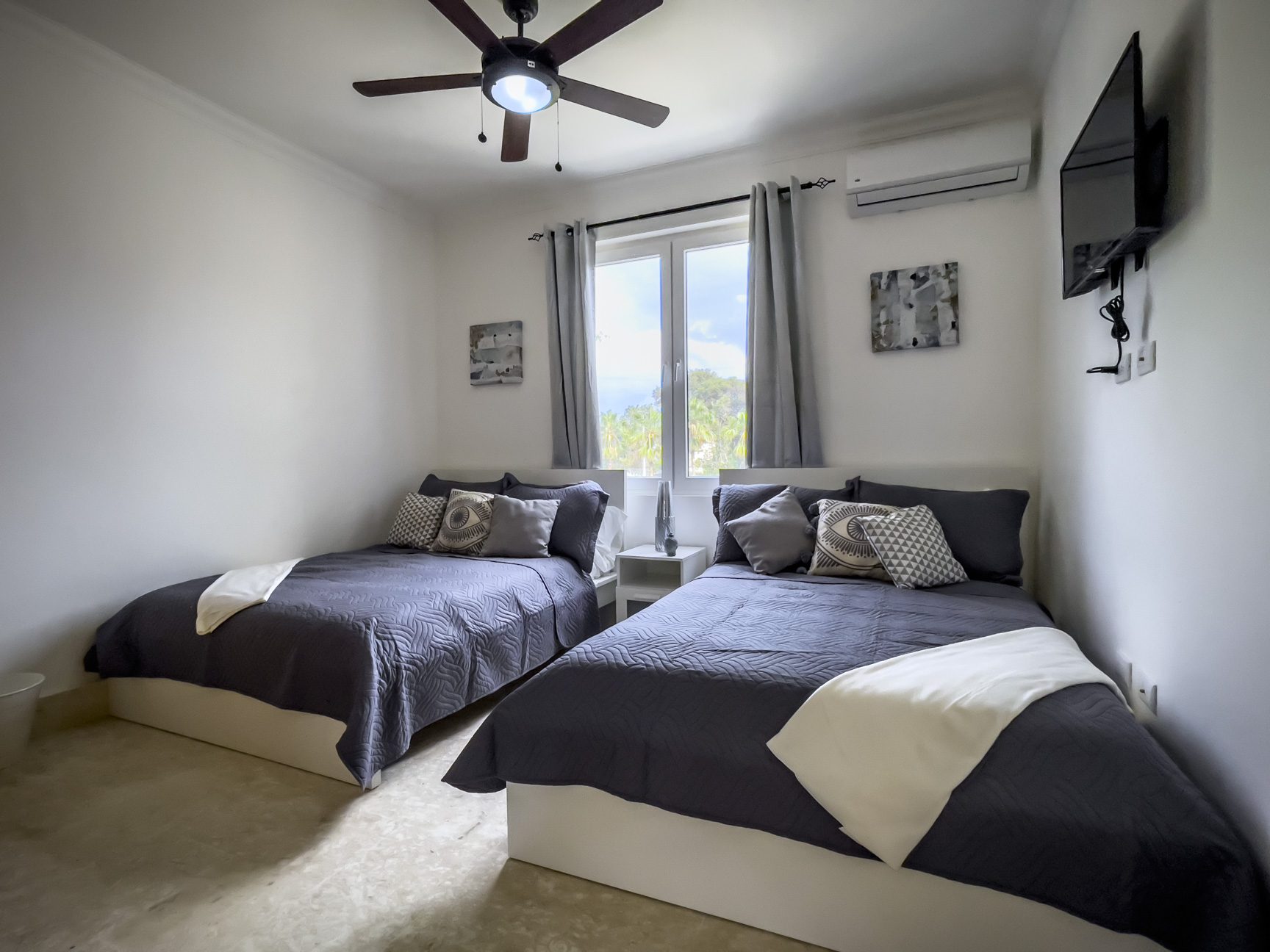buyDRproperty Bedroom with two beds, gray bedding, ceiling fan, and wall-mounted TV. Neutral decor. Sosua Real Estate