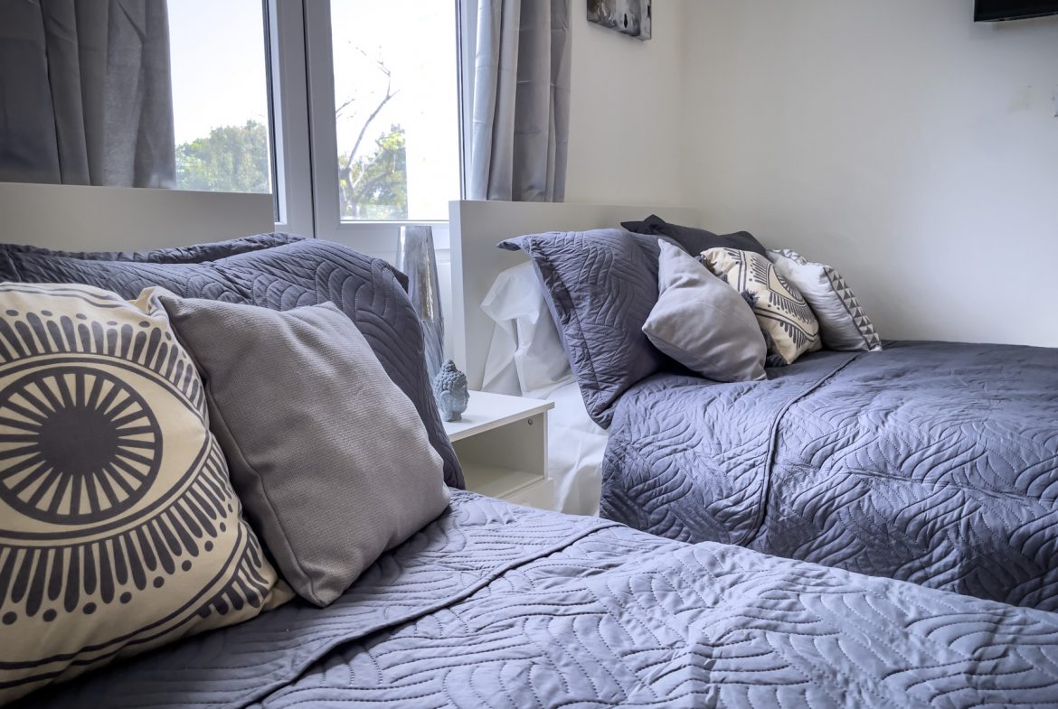 buyDRproperty Two twin beds with grey quilts and decorative pillows, bright window. Sosua Real Estate