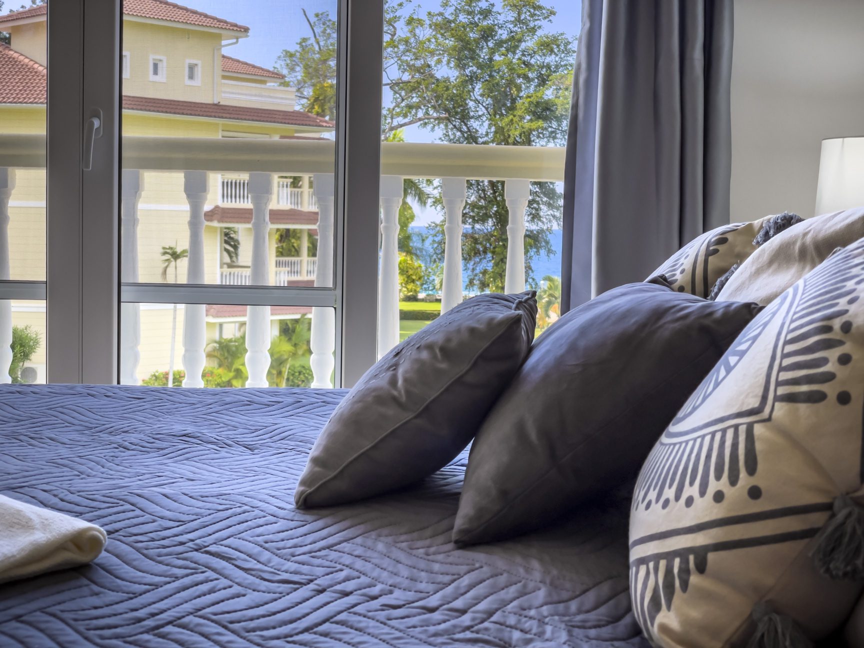 buyDRproperty Bright bedroom with a view of a tropical resort, featuring stylish pillows and a cozy blue quilt. Sosua Real Estate