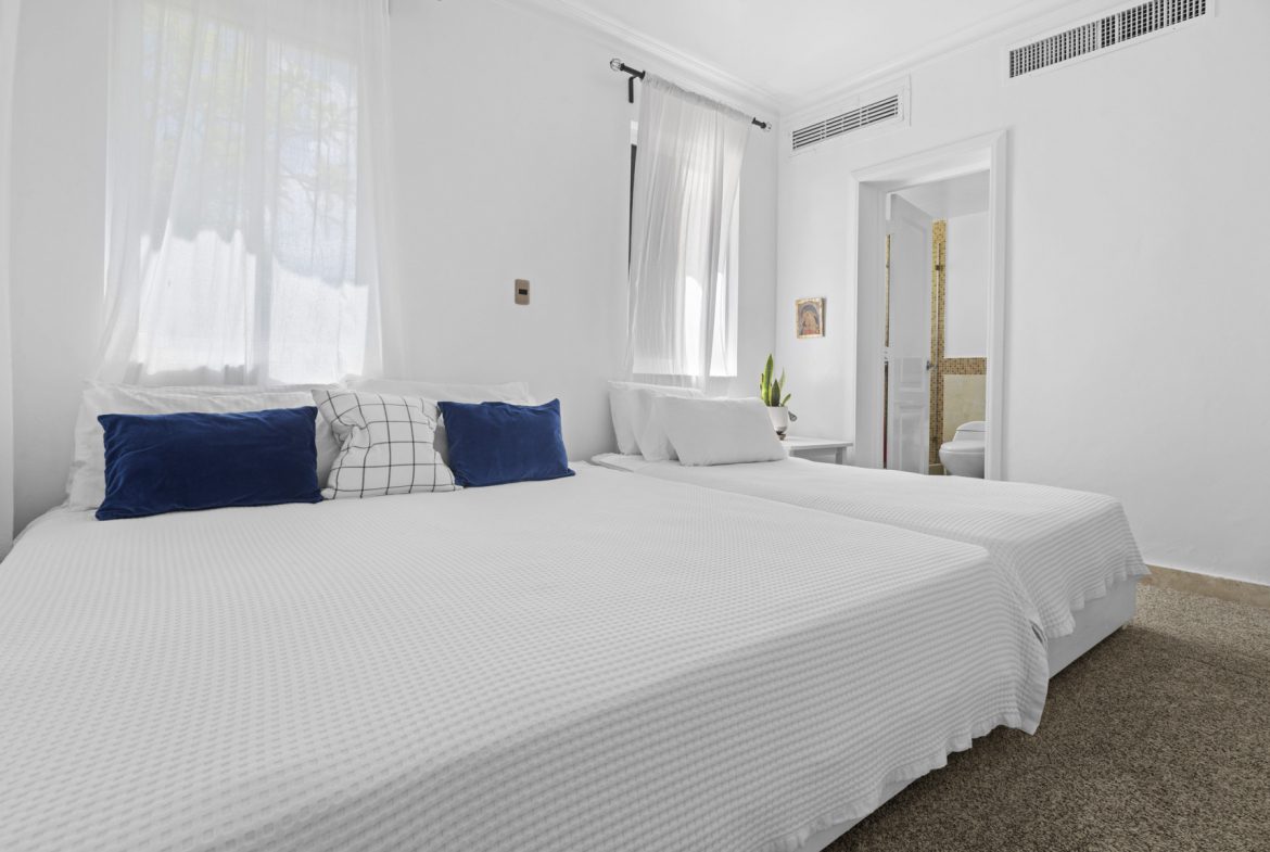 buyDRproperty Bright bedroom with two beds, white linens, blue pillows, and an ensuite bathroom. Sosua Real Estate