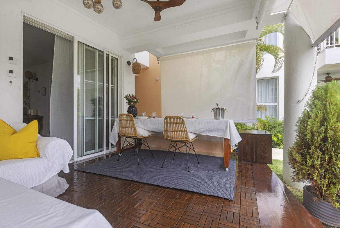 buyDRproperty Outdoor dining area with table set for a meal on a wooden patio, featuring a white sofa and lush greenery. Sosua Real Estate