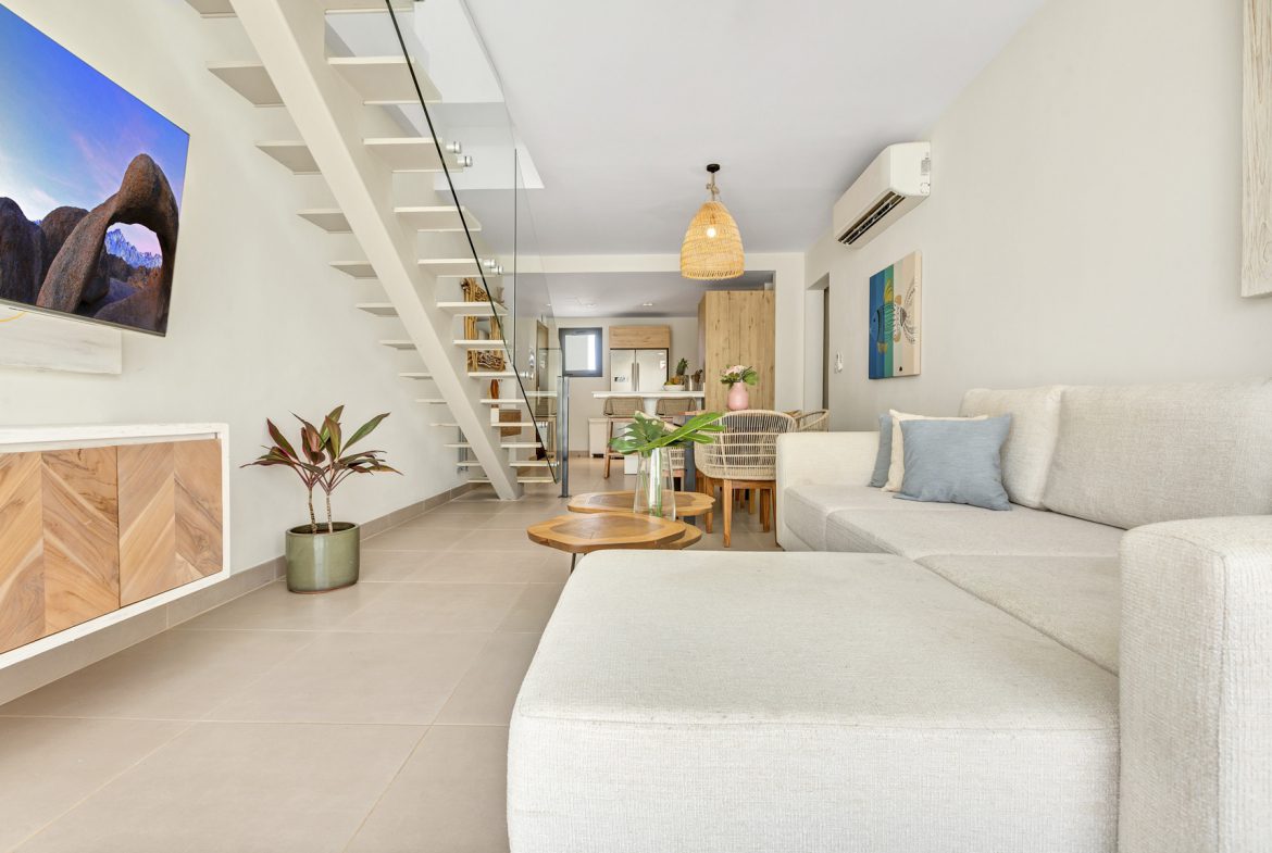 buyDRproperty Bright, modern living room with white sofa, wooden coffee tables, and staircase leading upstairs. Sosua Real Estate