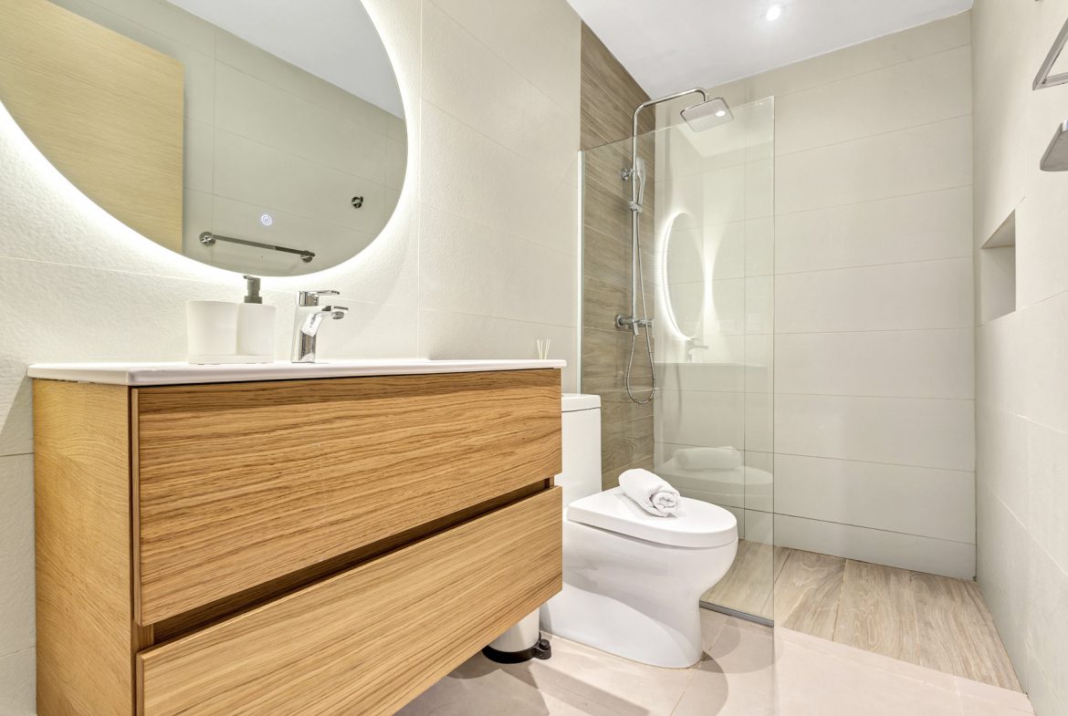 buyDRproperty Modern bathroom with wood vanity, round lighted mirror, glass shower, and white toilet. Sosua Real Estate