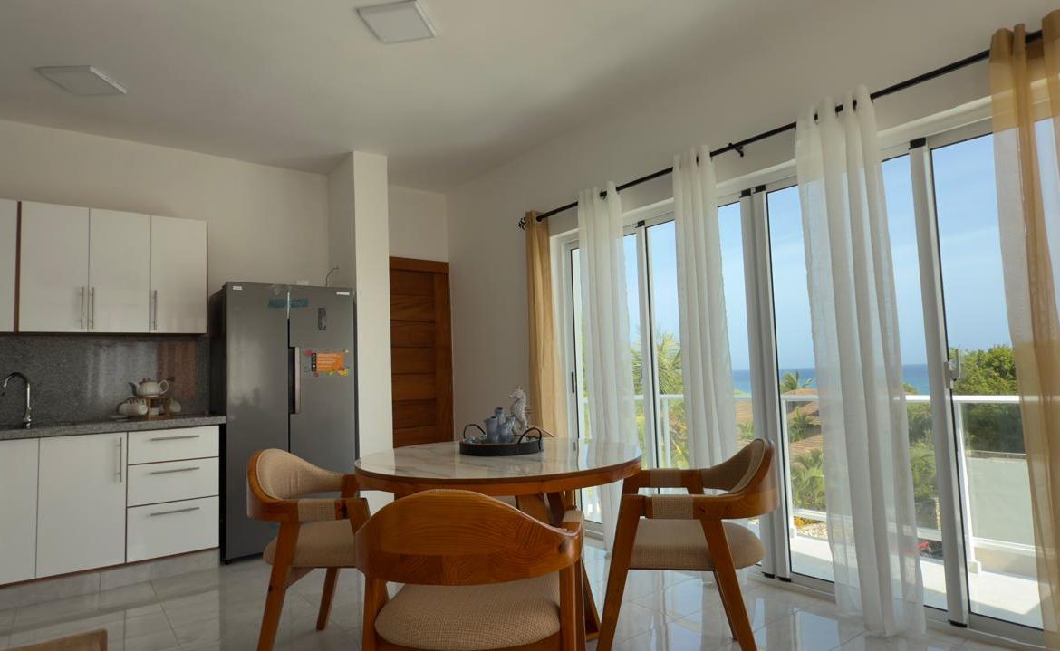 Bright dining area with round table, chairs, and large windows with ocean view.
