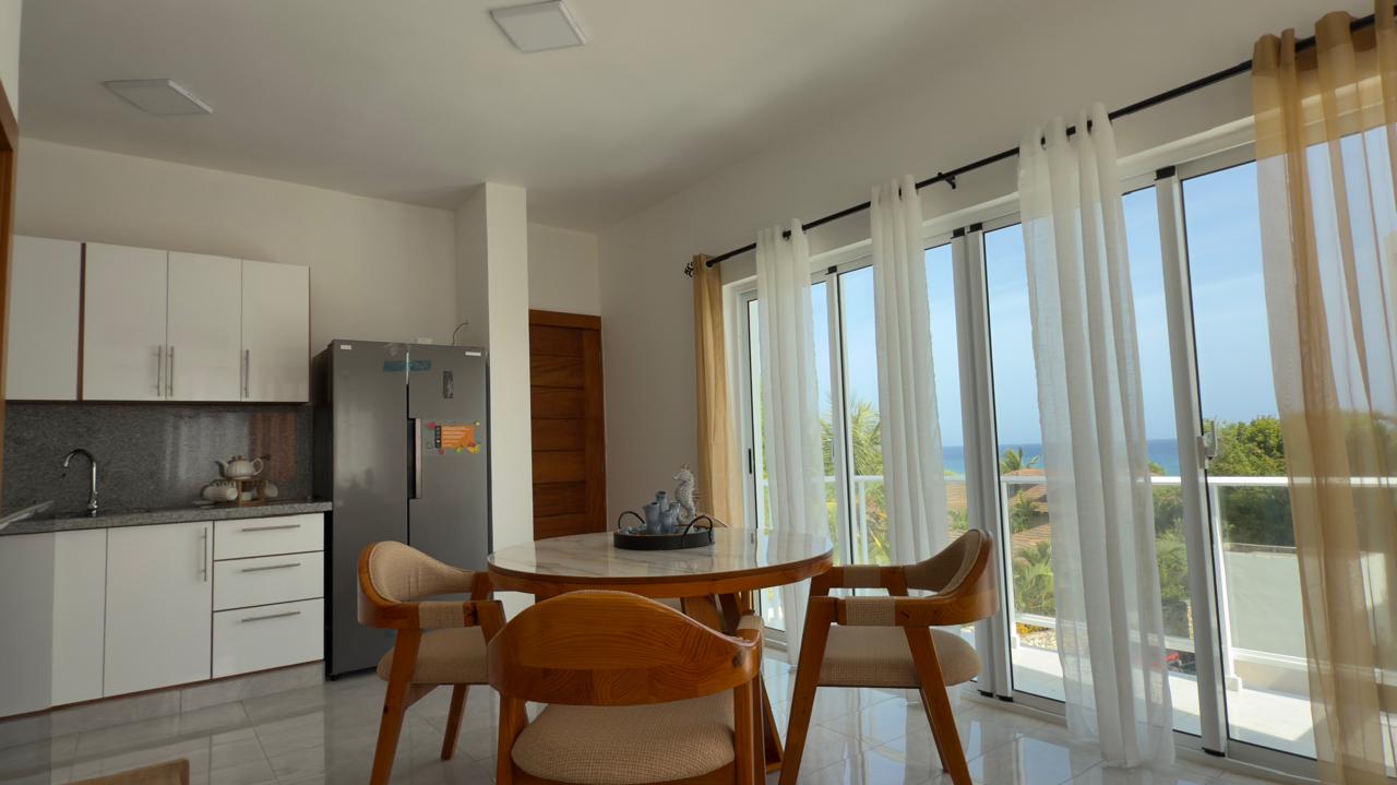 Bright dining area with round table, chairs, and large windows with ocean view.