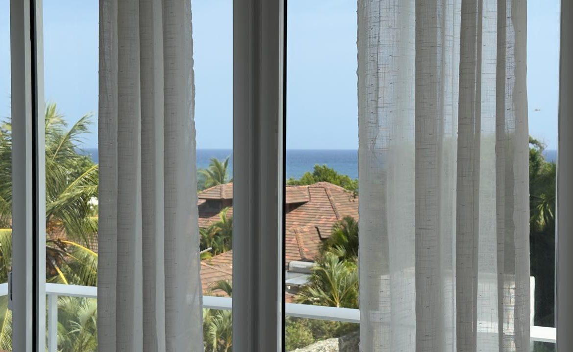 Ocean view from a window with white curtains, tropical trees, and rooftops.
