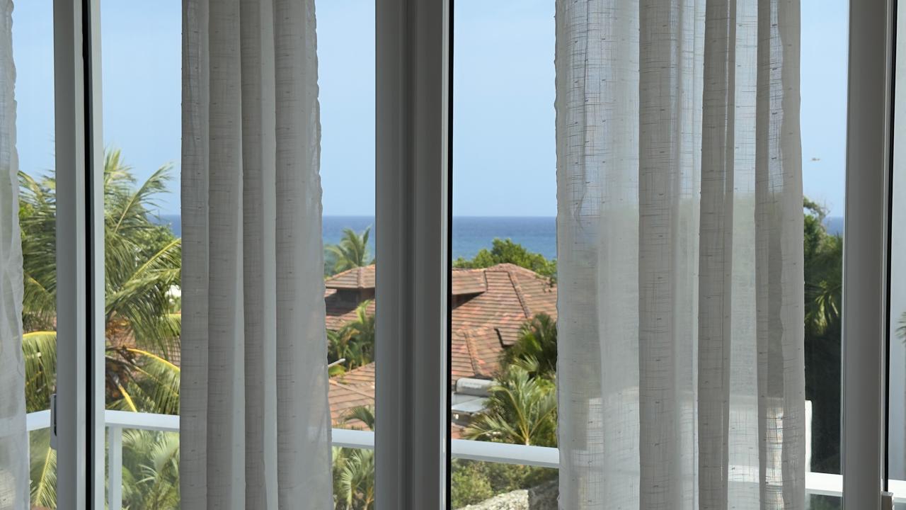Ocean view from a window with white curtains, tropical trees, and rooftops.