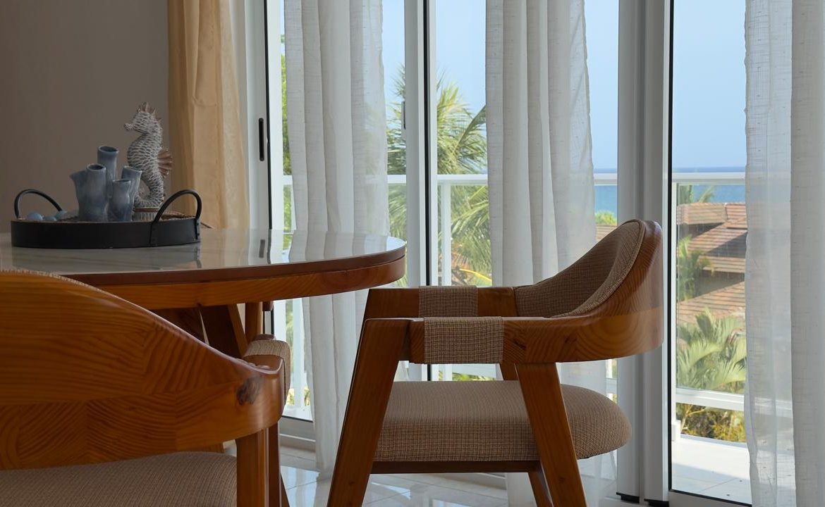Dining area with wooden chairs, glass table, and ocean view through sheer curtains.